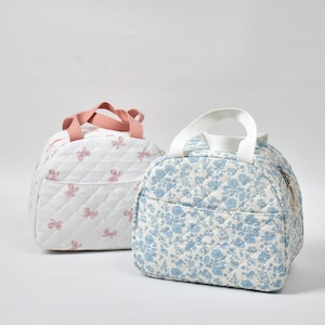 May include: Two quilted lunch bags. One is white with pink bow accents and a pink handle. The other is light blue with a floral pattern and a white handle. Both bags have a zippered pocket and a quilted design.