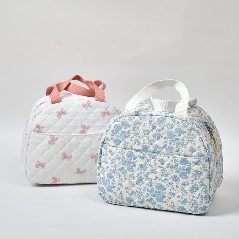 May include: Two quilted lunch bags. One is white with pink bow accents and a pink handle. The other is light blue with a floral pattern and a white handle. Both bags have a zippered pocket and a quilted design.