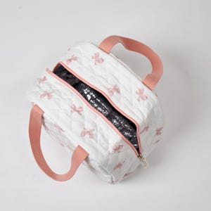 May include: White quilted lunch bag with pink bow accents and matching pink handles. The bag has a zipper closure and a black insulated interior. The bag is designed to keep food and drinks cool.