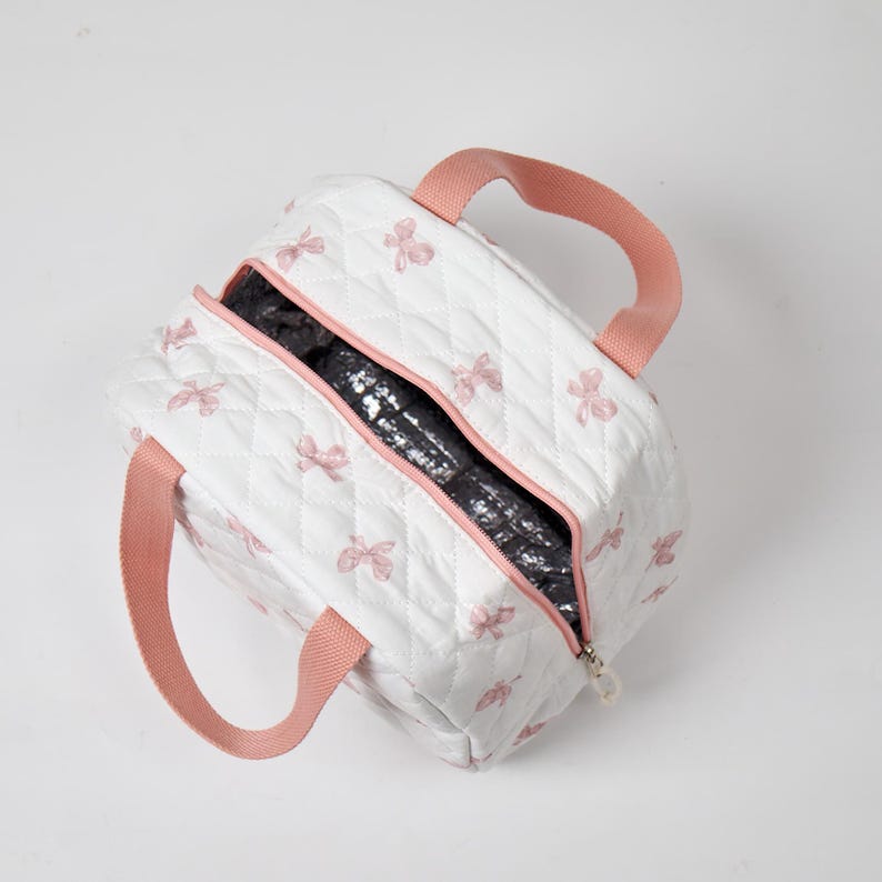 May include: White quilted lunch bag with pink bow accents and matching pink handles. The bag has a zipper closure and a black insulated interior. The bag is designed to keep food and drinks cool.