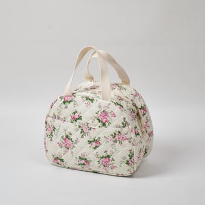 May include: A quilted, off-white bag with a floral pattern of pink roses and green leaves. The bag has a rounded shape, a zippered top, a front pocket, and two cream-colored handles. The bag is designed for carrying items.