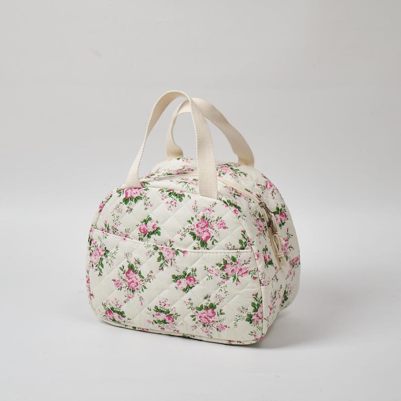 May include: A quilted, off-white bag with a floral pattern of pink roses and green leaves. The bag has a rounded shape, a zippered top, a front pocket, and two cream-colored handles. The bag is designed for carrying items.