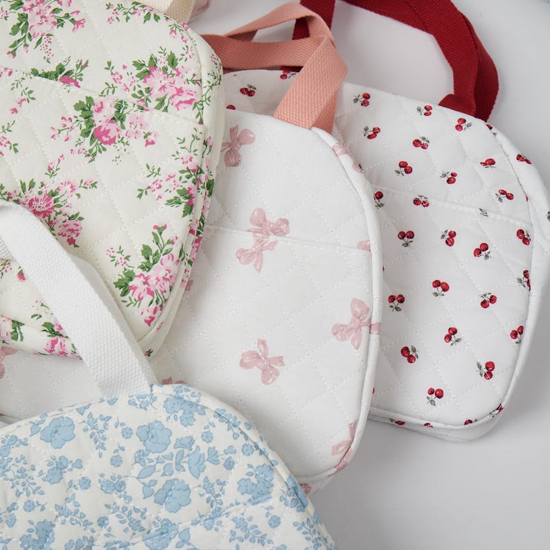 May include: A collection of quilted fabric bags with various floral and bow patterns. The bags feature white, pink, and blue fabrics with coordinating handles. The designs include pink roses, bows, and red cherries.