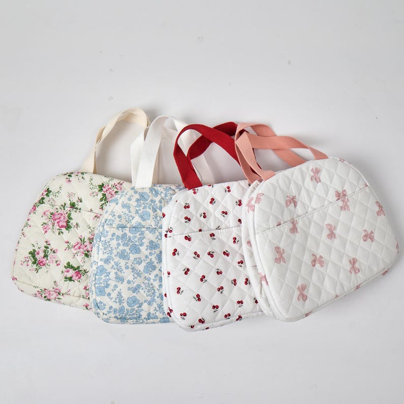 May include: Four quilted tote bags with different patterns and handle colors. The bags feature floral, cherry, and bow designs. The handles are cream, white, red, and pink. The bags have a quilted texture and a pocket on the front.
