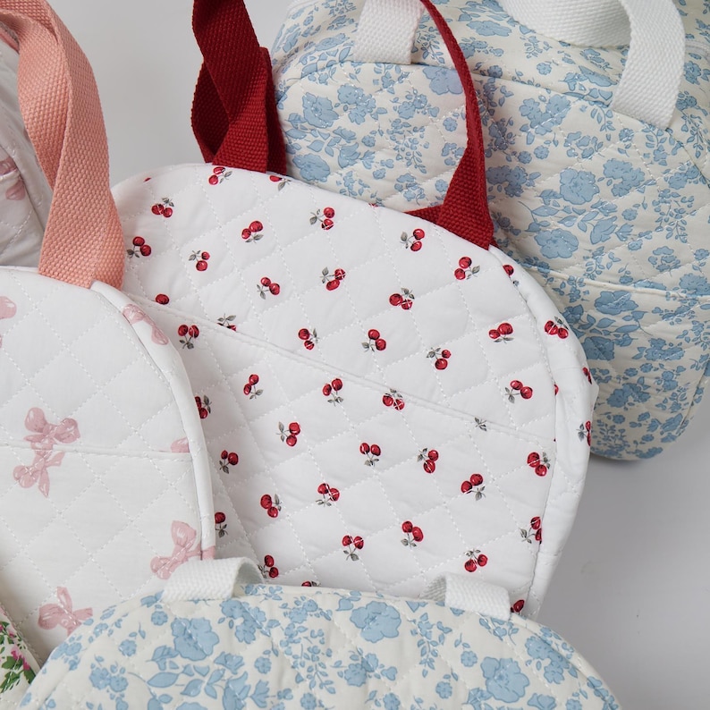 May include: Several quilted fabric tote bags in various patterns. One bag features a white background with red cherries, another with pink bows, and a third with blue floral designs. The bags have different colored handles.