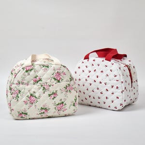 May include: Two quilted lunch bags. One is cream-colored with a pink floral pattern, a cream handle, and a front pocket. The other is white with a red cherry pattern and a red handle. Both bags have a zippered closure.