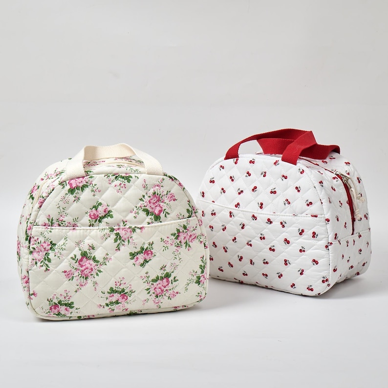 May include: Two quilted lunch bags. One is cream-colored with a pink floral pattern, a cream handle, and a front pocket. The other is white with a red cherry pattern and a red handle. Both bags have a zippered closure.