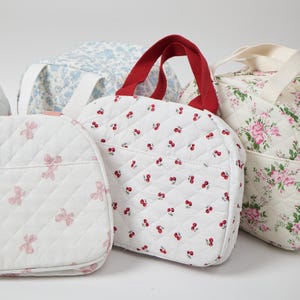 May include: Several quilted tote bags in various floral and fruit patterns. One bag features a white background with red cherries, another with pink bows, and others with blue and pink floral designs. The bags have red and cream-colored handles.