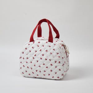 May include: White quilted bag with a cherry pattern and red handles. The bag has a rounded shape, a zipper closure, and a small pocket on the front. The handles are a deep red color.
