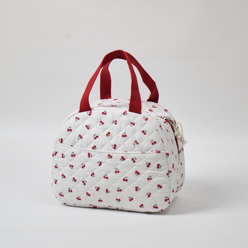 May include: White quilted bag with a cherry pattern and red handles. The bag has a rounded shape, a zipper closure, and a small pocket on the front. The handles are a deep red color.