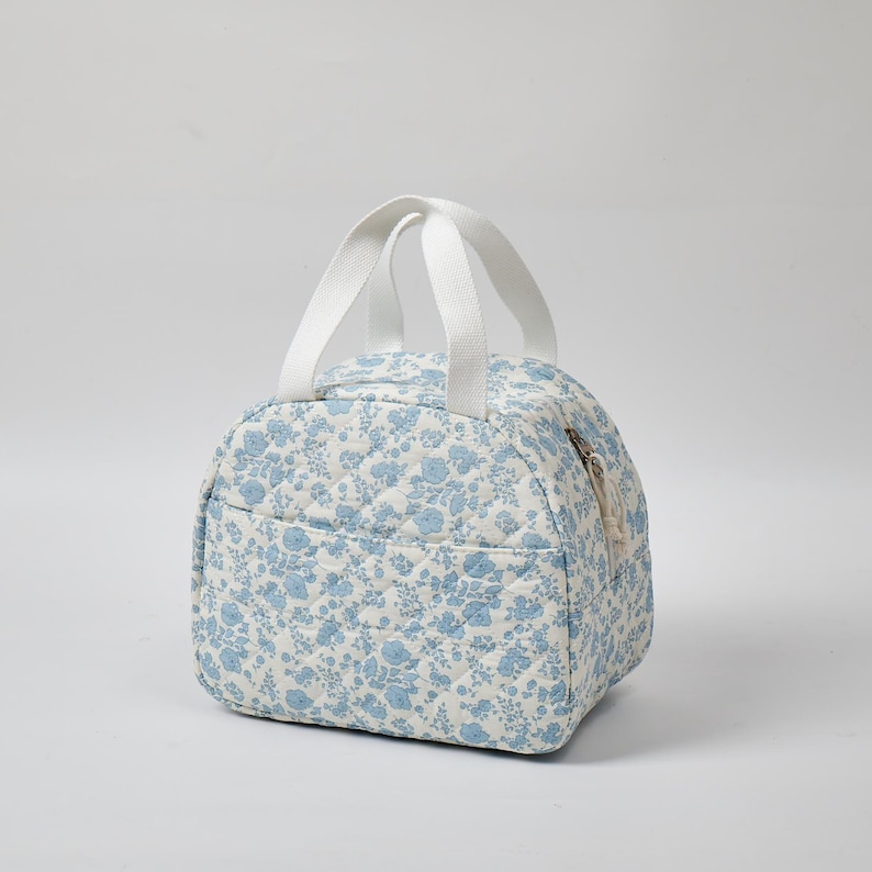 May include: A quilted lunch bag with a floral pattern in light blue and white. The bag has a zippered closure, a small front pocket, and two white handles. The bag is approximately 8 inches wide and 6 inches tall.