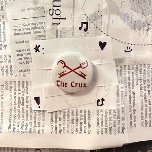 以下が含まれることがあります： 赤色の太字で「The Crux」と書かれた丸いオフホワイトのボタン。ボタンには、赤い交差したシャベルと鍵のデザインが施されています。ボタンは、新聞の切り抜きの背景に配置されています。