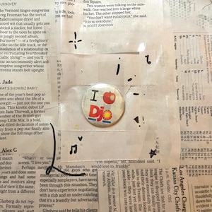 May include: A circular badge with the words "I DJ" and an apple illustration. The badge is red, white, and blue, and is placed on a background of newspaper print.