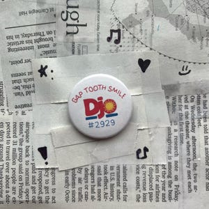 以下が含まれることがあります： 赤と青で「GAP TOOTH SMILE」の文字と太陽のグラフィックが入った白いボタン。 下には「Djo」と番号「#2929」があります。 ボタンは新聞の切り抜きの背景にあります。