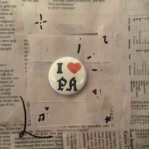 Puede incluir: Un botón blanco con el texto "I PA" y un corazón rojo. El botón está sobre un fondo de recortes de periódico y cinta adhesiva.