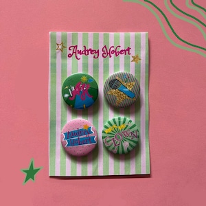 Op de afbeelding: Een set van vier ronde buttons bevestigd op een lichtgroen en wit gestreepte displaykaart met de tekst Audrey Hobert. De buttons bevatten kleurrijke ontwerpen waaronder een microfoon, de naam Audrey Hobert en de tekst Who's the Clown.