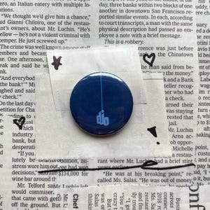 May include: A navy blue button with a white graphic and the letters "dio". The round button is placed on a newspaper background with handwritten notes and drawings.