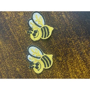 May include: Two embroidered bee patches. Each patch features a yellow bee with black stripes, white and yellow wings, and a smiling face. The patches are on a brown, textured surface.