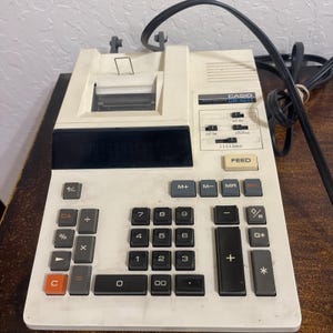 Op de afbeelding: Een vintage Casio DR-1011 rekenmachine met ingebouwde printer. De rekenmachine is gebroken wit met zwarte en grijze toetsen, waaronder een numeriek toetsenbord, functietoetsen en een "FEED"-knop. Een zwart netsnoer is bevestigd.
