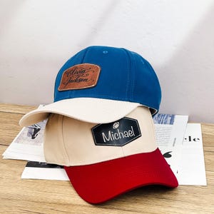 Puede incluir: Una gorra de béisbol azul con visera beige y un parche de cuero marrón con el nombre Olivia Jackson, sobre una gorra beige con visera roja y un parche hexagonal negro con un icono de balón de fútbol y el nombre Michael en blanco.