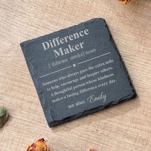 May include: A square dark gray slate coaster with rough, uneven edges. The surface is engraved with the text: Difference Maker, followed by a phonetic pronunciation, a definition of the term, and the name Emily at the bottom.