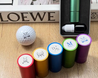 Könnte beinhalten: Ein Golfball und mehrere zylindrische Golfaccessoires in verschiedenen Farben, darunter Rot, Gold, Blau, Grün und Lila. Jeder Zylinder hat ein anderes Logo auf der Oberseite. Ein grünes Golfzubehör befindet sich in einem Metallgehäuse.