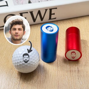 May include: A white golf ball with a personalized face design, next to a blue and a red cylindrical container, each with a matching face design. The items are on a wooden surface.