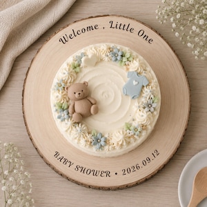May include: A round wooden serving board engraved with "Welcome, Little One" and "BABY SHOWER 2026.09.12" featuring a white frosted cake decorated with a brown teddy bear, a light blue baby onesie, a white heart, and light blue and green flowers.