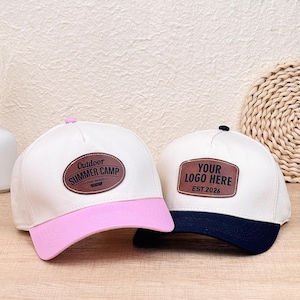 Può includere: Due cappellini da baseball con corona bianca e toppe in similpelle marrone. Il cappellino a sinistra ha una visiera rosa e una toppa con la scritta Outdoor SUMMER CAMP PINE WOOD. Quello a destra ha una visiera blu navy e YOUR LOGO HERE EST.2026.