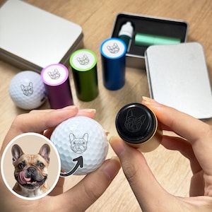 May include: A golf ball with a French bulldog design is held next to a stamp with the same design. Several colored stampers and a small metal container are in the background. The stampers are purple, green, and blue.