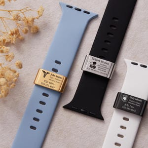 Puede incluir: Tres correas de Apple Watch en azul claro, negro y blanco. Cada correa presenta una etiqueta de identificación médica de metal con texto grabado. La correa azul claro tiene una etiqueta dorada con un símbolo médico y el nombre "John Smith". Las correas negras y blancas tienen etiquetas plateadas y negras con nombres e información médica.