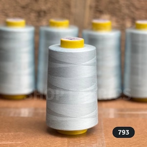 May include: A collection of light grey thread spools with yellow tops, set against a brown backdrop. The thread is neatly wound, and the number 793 is displayed in a black oval on the right.