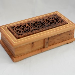 Könnte beinhalten: Eine rechteckige Holzbox mit einem geschnitzten Blumendesign auf dem Deckel. Die Box ist hellbraun mit einer dunkelbraunen, aufwendig geschnitzten Platte oben. Der Deckel hat einen dünnen, hellfarbigen Rand. Die Box steht auf weißem Hintergrund.