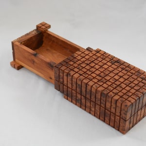 Puede incluir: Una caja de madera con tapa corredera. La caja es de madera marrón claro y tiene forma rectangular. La tapa está compuesta por pequeños bloques cuadrados, creando una superficie texturizada. La caja está abierta, revelando el interior.