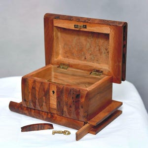 Puede incluir: Una caja rectangular de madera con la tapa abatible abierta, que presenta un patrón de veta de madera marrón natural. El panel frontal tiene ranuras verticales talladas y una cerradura. Una pequeña llave de latón y una pieza de madera suelta yacen frente a la caja.