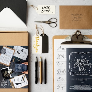 May include: An assortment of calligraphy supplies, including a brush calligraphy kit, pens, tags with "thank you" and "with love" text, scissors, envelopes, and a clipboard with calligraphy practice sheets.