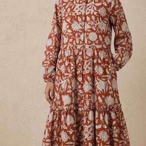 May include: A long-sleeved, button-down dress in a rust color, adorned with a white floral pattern. The dress features a collared neckline, tiered skirt, and falls to mid-calf length. The model is wearing brown sandals.