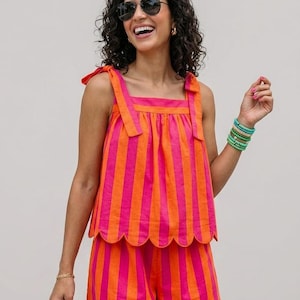 Op de afbeelding: Een tweedelige set bestaande uit een mouwloze top en een short, beide voorzien van verticale felroze en oranje strepen. De top heeft schouderbandjes om te strikken en een geschulpte zoom, terwijl de bijpassende short ook een geschulpte zoom heeft.
