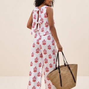 May include: A white sleeveless top and matching wide-leg trousers featuring a repeating red and pink floral block print. The top has a back cutout with two tied bows. A large, tan woven straw tote bag with dark brown leather straps is held at the side.