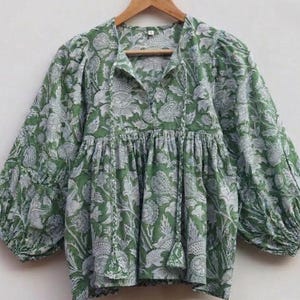 May include: A green and white floral print blouse featuring a V-neckline with long braided ties ending in tassels. The garment has voluminous long sleeves with gathered cuffs and a pleated bodice that creates a loose, flowing silhouette.