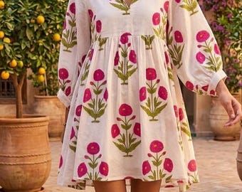 May include: White mini dress with a pink floral print and green stems. The dress has long sleeves and a V-neck. A straw hat and gold necklaces complete the look. The background features a courtyard with plants and a water feature.