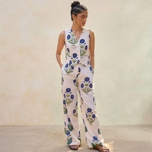 May include: A two-piece outfit comprising a sleeveless top and wide-leg trousers. The white fabric features a repeating pattern of blue flowers and green leaves. The outfit is worn with brown sandals.