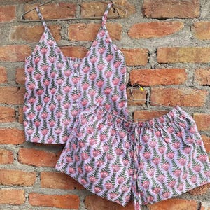 May include: A two-piece pajama set consisting of a sleeveless camisole top with thin straps and a button-down front, paired with matching elastic-waist shorts. Both pieces feature a repeating pattern of pink flowers with green leaves on a light purple background.