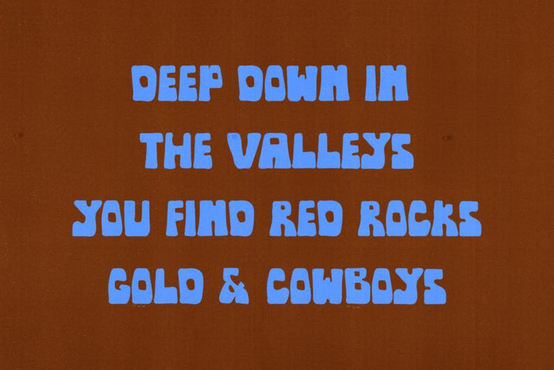 May include: A brown background with the blue text: "DEEP DOWN IN THE VALLEYS YOU FIND RED ROCKS GOLD & COWBOYS". The text is in a bold, rounded font. The overall design has a vintage, retro aesthetic.