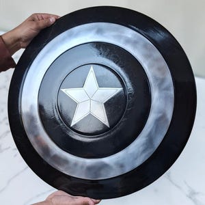 Op de afbeelding: Een zwart en zilver schild met een witte ster in het midden. Het schild is rond met concentrische cirkels van zwart en zilver. De ster is een vijfpuntige ster. Het schild wordt door twee handen vastgehouden.