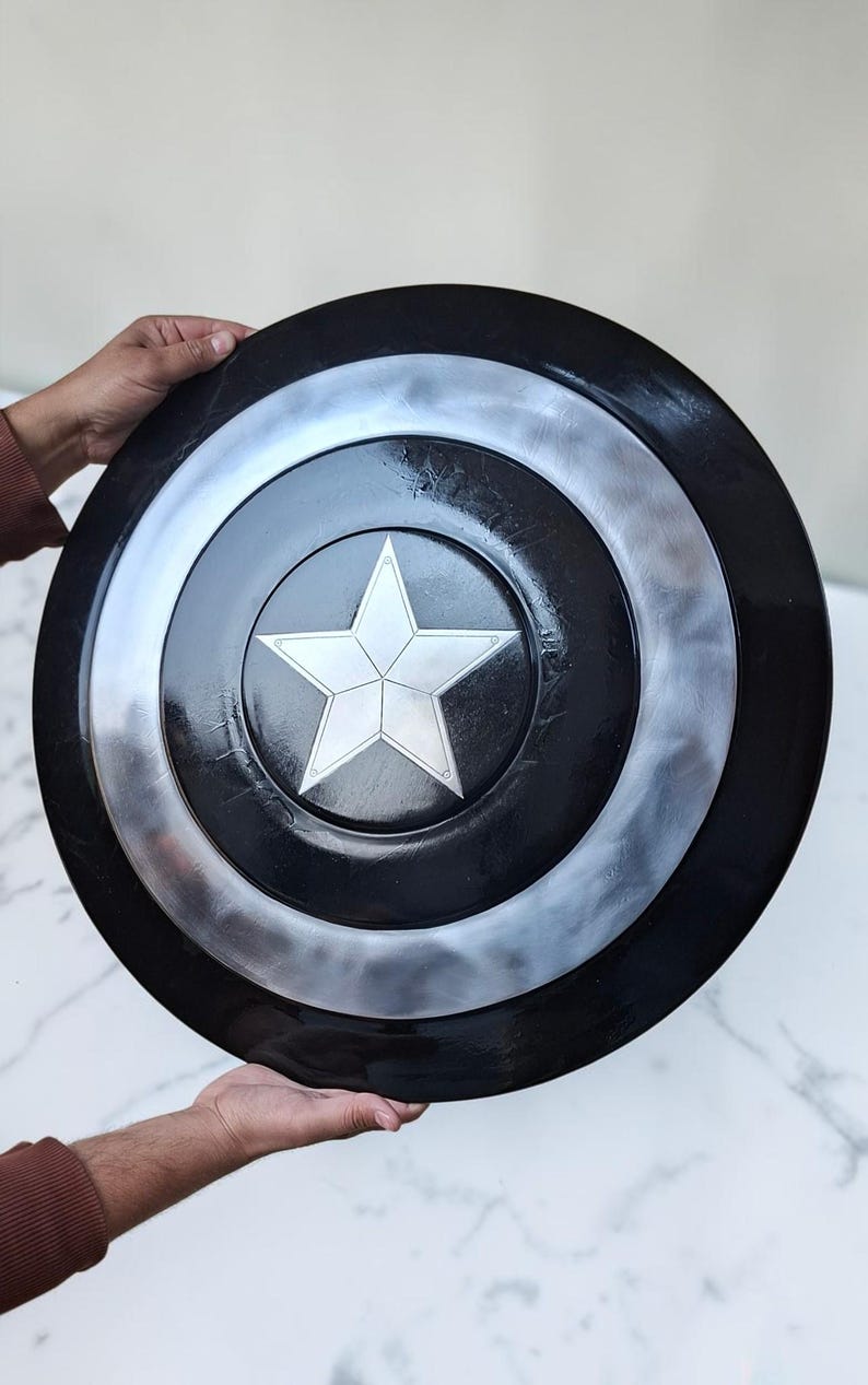 Op de afbeelding: Een zwart en zilver schild met een witte ster in het midden. Het schild is rond met concentrische cirkels van zwart en zilver. De ster is een vijfpuntige ster. Het schild wordt door twee handen vastgehouden.