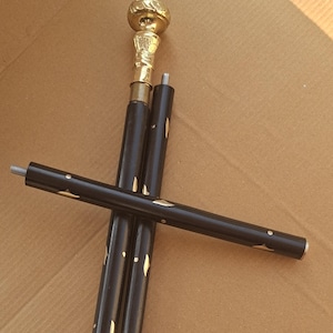 Puede incluir: Un bastón de madera negro con un mango y detalles dorados. El mango tiene un diseño detallado y ornamentado. El bastón está desmontado, con las piezas sobre una superficie marrón.
