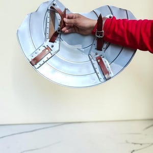 May include: A silver-colored shield with brown leather straps and rivets. The shield has a curved shape and is held by a person wearing a red shirt. The shield is likely for cosplay or historical reenactment.