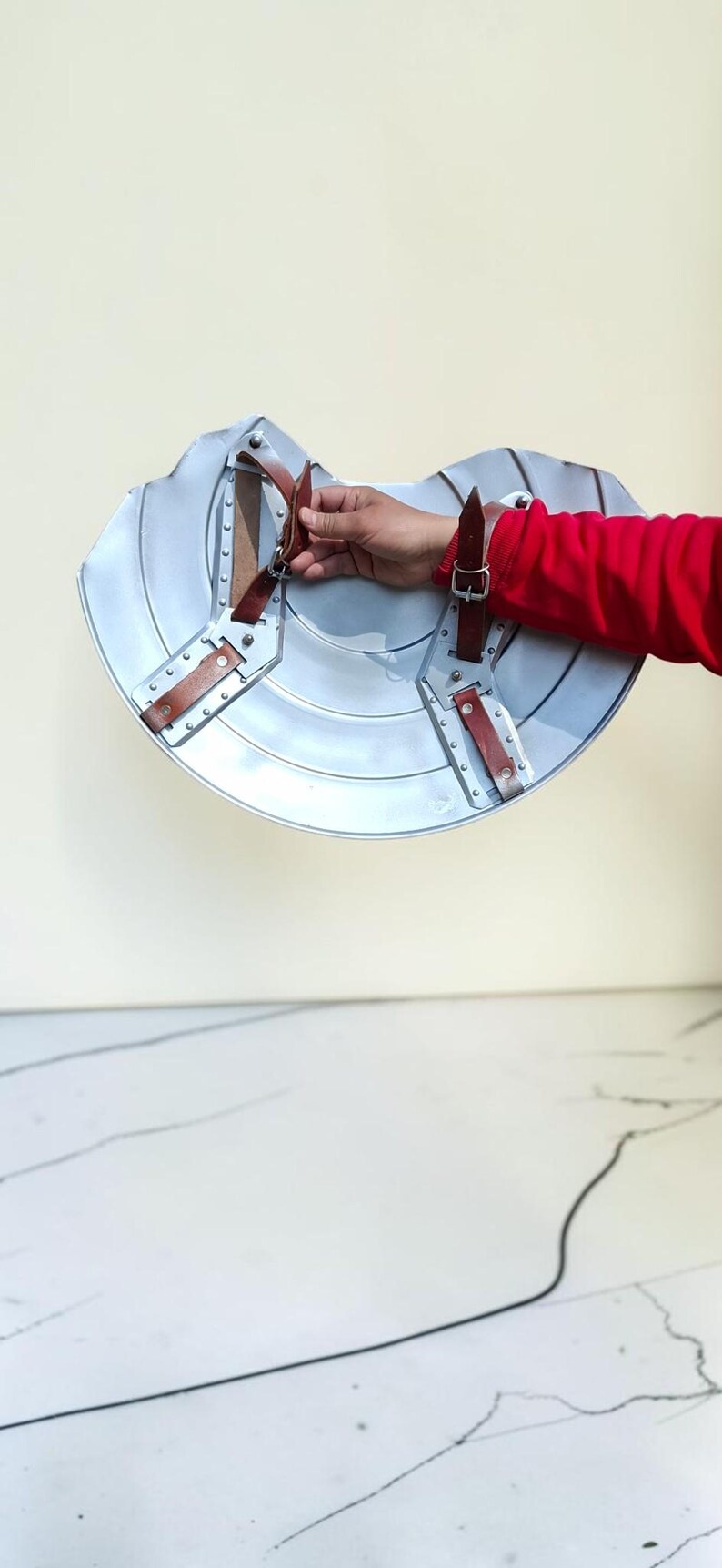 May include: A silver-colored shield with brown leather straps and rivets. The shield has a curved shape and is held by a person wearing a red shirt. The shield is likely for cosplay or historical reenactment.