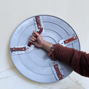 Op de afbeelding: Een zilverkleurig, cirkelvormig schild met een reeks concentrische groeven. Het schild heeft vier bruine leren riemen die met zilveren klinknagels zijn bevestigd. Het schild wordt door een persoon vastgehouden.